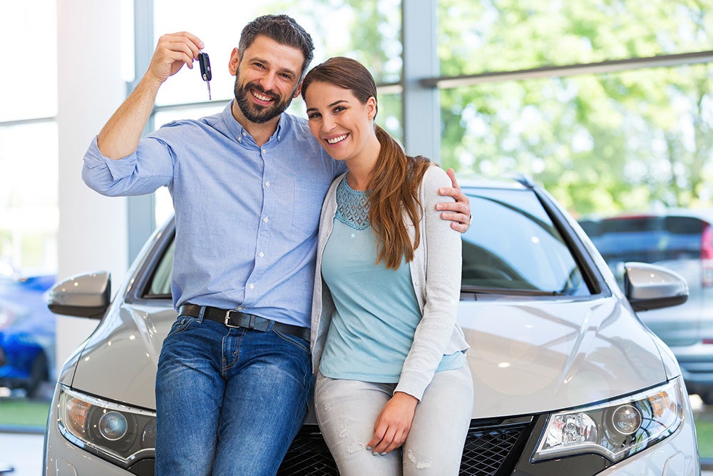 Smiling couple leaning on a new car in a dealership, man holding up car keys happily.
